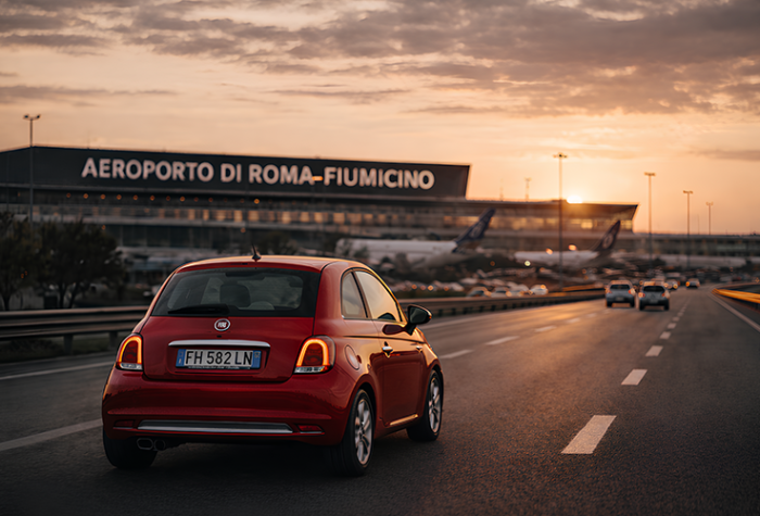 Rental car driving away from Rome Fiumicino Airport at sunset, showing the terminal building and road access toward the city and surrounding regions.