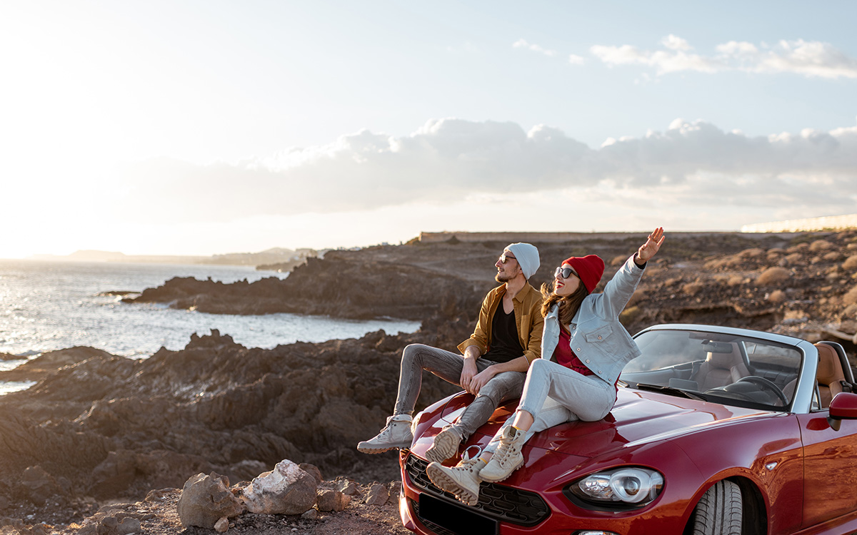 Giovane coppia innamorata che ammira il paesaggio, seduta insieme sul cofano dell’auto lungo una costa rocciosa sull’oceano. Atmosfera spensierata, amore e libertà di viaggiare.