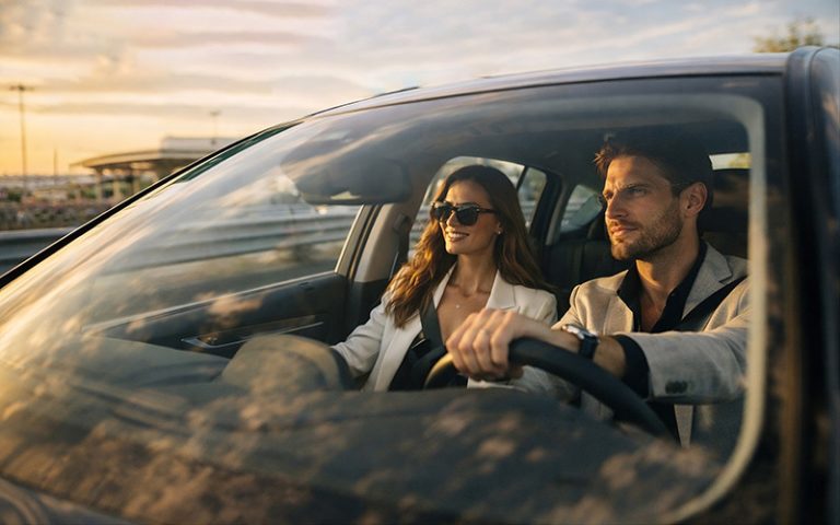 Stylový pár řídí pronajaté auto při západu slunce, což znázorňuje typickou cestovatelskou situaci po rezervaci vozidla s využitím alternativních platebních metod.