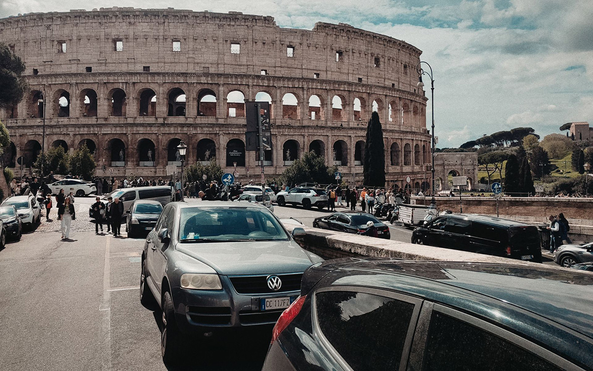 Cars driving and parking near the Colosseum in Rome with historic architecture in the background.
