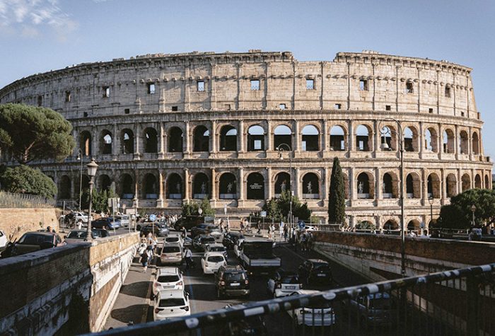 Driving in Rome near the Colosseum with busy traffic, cars, and pedestrians around one of the city's most famous landmarks.