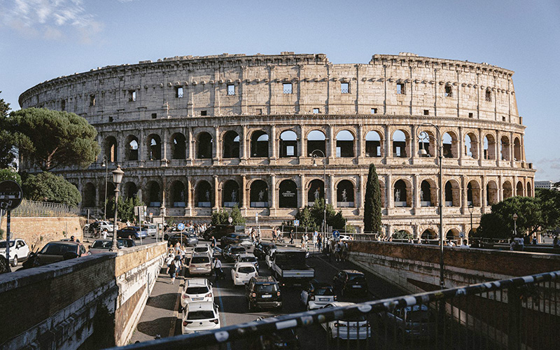 Driving in Rome near the Colosseum with busy traffic, cars, and pedestrians around one of the city's most famous landmarks.