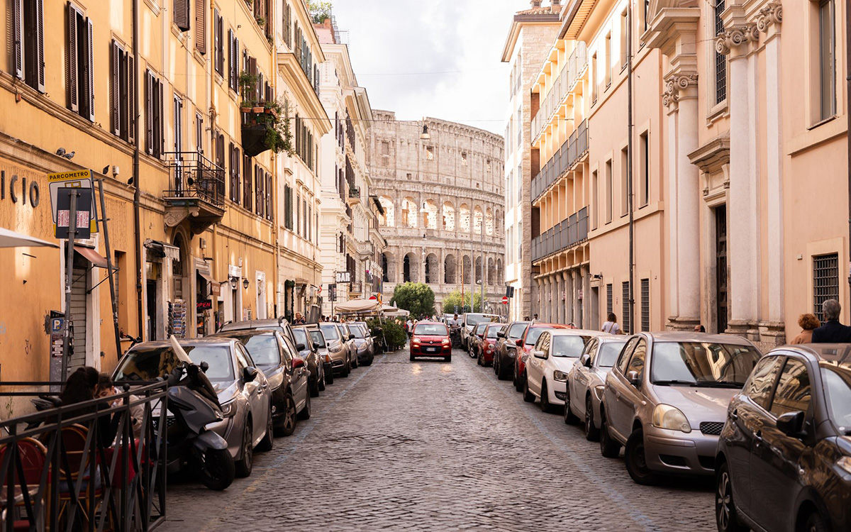 Narrow cobblestone street in Rome with parked cars and the Colosseum visible in the background.